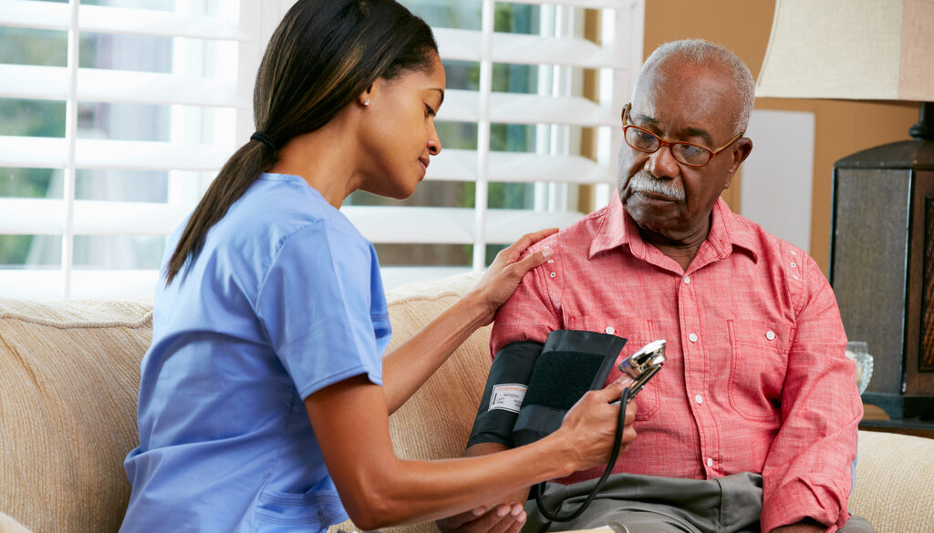 African American Nurse Checking an Elderly Black Man’s Blood Pressure on the Couch at Home in Home Health Care