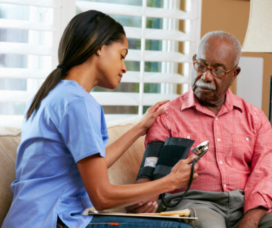 African American Nurse Checking an Elderly Black Man’s Blood Pressure on the Couch at Home in Home Health Care